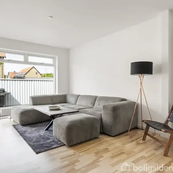 Gråt sofaarrangement står i moderne stue med trægulv, ved siden af er en sort gulvlampe og stol. Store vinduer åbner til balkon med udsigt til træer og huse.
