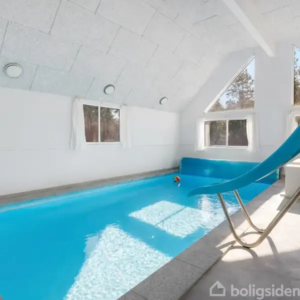 Indendørs swimmingpool med blå vandrutsjebane, placeret i et lyst rum med hvide vægge og vinduer, der lukker naturligt lys ind og afslører en udendørs skovscene.