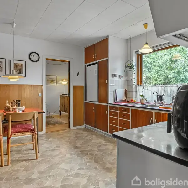 Et køkken med retro træmøbler har en spiseplads med to stole og et bord dækket med frugt. Store vinduer giver udsigt til grønne træer, og der hænger lamper over bordet.