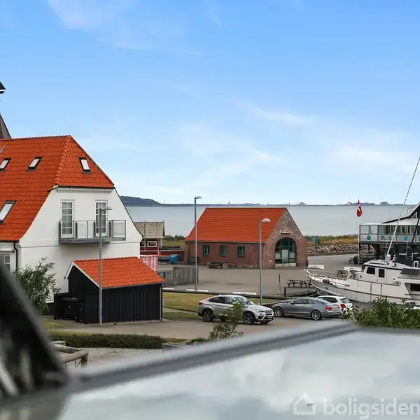 Røde tegltage på hvide bygninger ved en havn. Både ligger fortøjet langs kajen i baggrunden, og himlen er blå med få skyer.