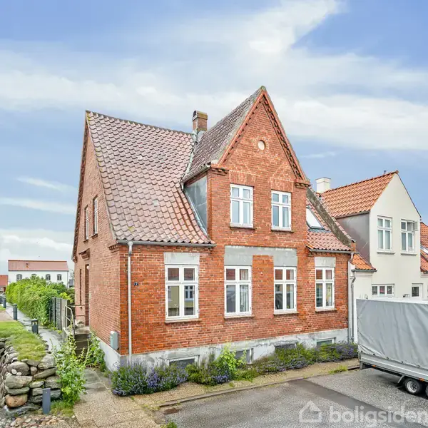 Et rødt murstenshus står stille i et boligkvarter. Ved siden af huset ses en overdækket trailer på en asfalteret vej. Husene omkring har også røde tage.