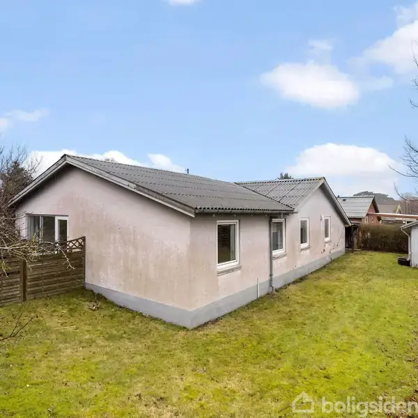 Et lyserødt hus står stille omgivet af en grøn græsplæne og buske. I baggrunden ses et lille hvidt skur, og himlen er blå med lette skyer.