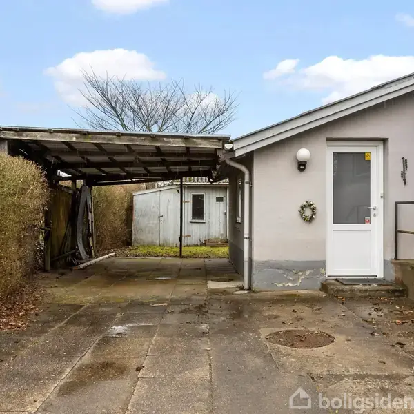 Hus med concret indkørsel leder til carport. Tiltstedeværelse af vinterhæk omkring området. Grå himmel og døren prydet med krans.