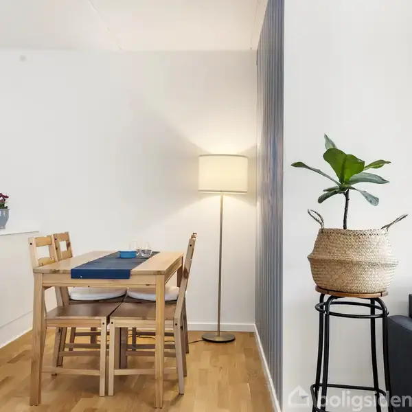 Træspisebord omgivet af fire stole har en blå bordløber på sig. Ved siden står en gulvlampe og en plante i en kurv. Væggene er hvide og gulvet er træ.