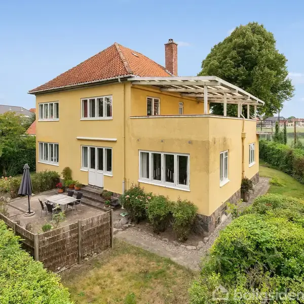 Et gult hus med rødt tegltag står stille i en grøn have med en terrasse. En hæk omkranser huset, og et åbent landskab ses i baggrunden.