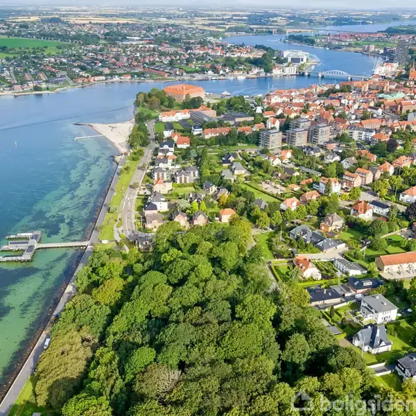 En by ligger ved siden af en stor vandmasse. Huse og bygninger strækker sig langs kysten, med en strand og bådebro. Skovområder omkranser byens udkant.