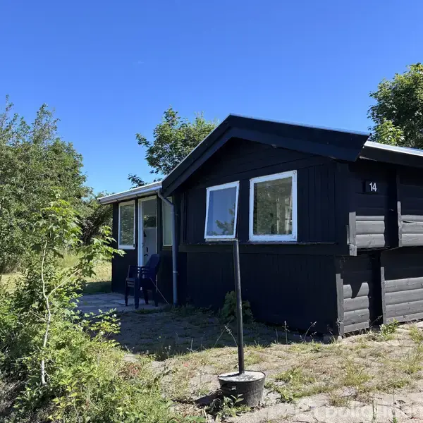 Sort træhus står stille i en grøn, solbeskinnet have. Nummeret "14" ses på husets gavl. Vegetation omgiver huset, og en blå himmel strækker sig ovenover.
