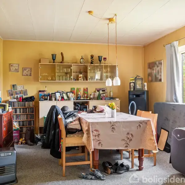 Et spisebord dækket med en dug står i en stue omgivet af stole, hylder med dekorative genstande og en reol med bøger og cd'er; vindue med gardiner til højre.