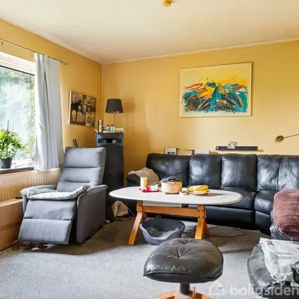 Stue med sort lædersofa og hvilestol placeret omkring et rundt sofabord. Maleri hænger på væggen. Klart dagslys strømmer ind gennem vinduet med gardiner, udsigt til have.
