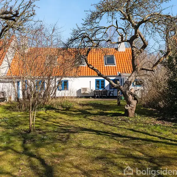 Hus med hvid facade og orange tegltag i baggrunden. Træer står i en grøn have. Bord med stole ses foran huset, omgivet af buske og træer.