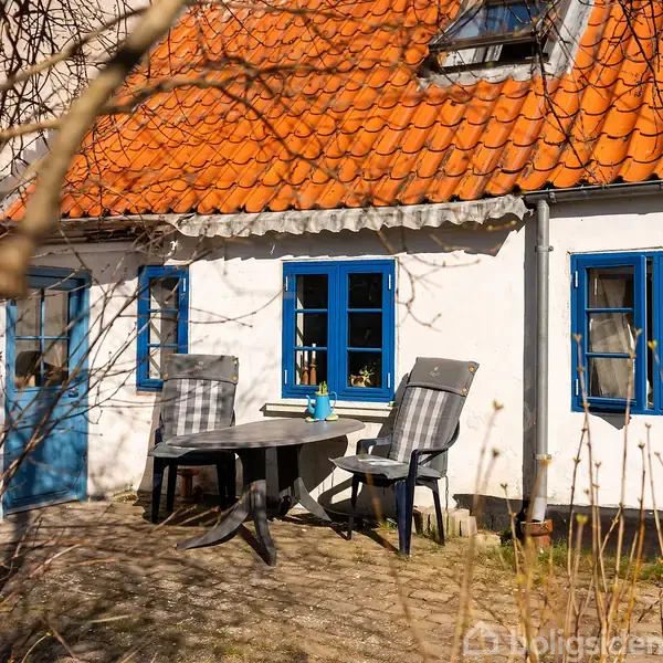 Huset har en hvid murstensfacade med blå vinduesrammer og en orange tegltag. To stole og et bord står på terrassen. Omgivelserne er præget af nøgne grene og buske.