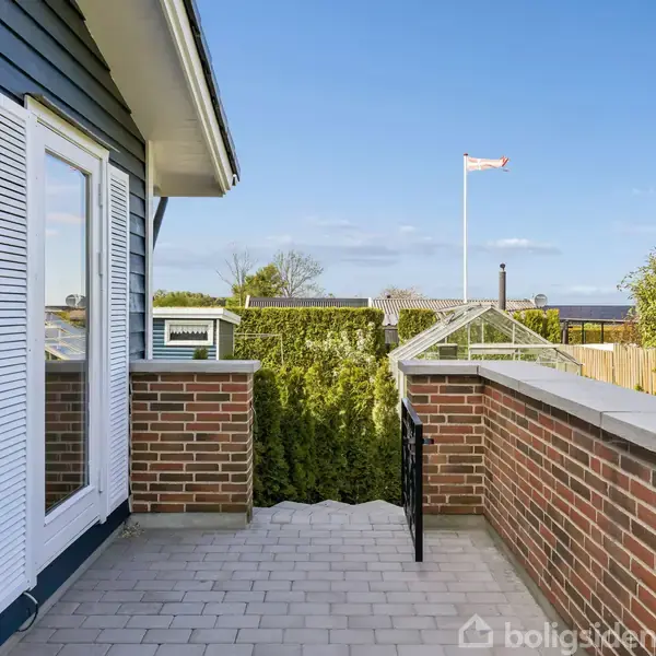 Terrasse med røde murstensmure, beliggende ved siden af et hus med blå træbeklædning og hvidt vindue foran en have med buske og træer under en blå himmel.
