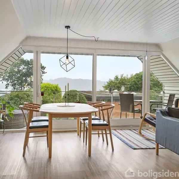 Et lyst træbord med stole står i en moderne stue med trægulv, omgivet af store vinduer, der fører ud til en terrasse med grønne planter og udsigt over landskabet.