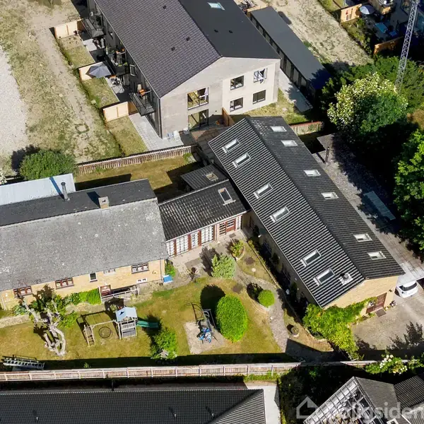 Boligkompleks set fra oven med to bygninger i vinkel omkring en velplejet have. Omgivelserne består af andre huse, grønne områder og en parkering.