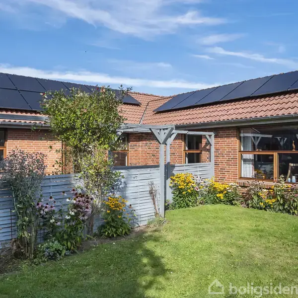 Rødt murstenshus med installerede solpaneler på taget står i en have med blomster og græsplæne. En lav hegnsstruktur adskiller huset fra haven.