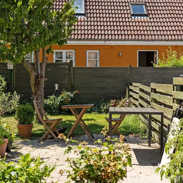 Havebord med to stole står på en flisebelagt terrasse i en have. Omkring er der grønne planter og buske. En hvidmalet husmur ses i baggrunden gennem et grønmalet hegn.