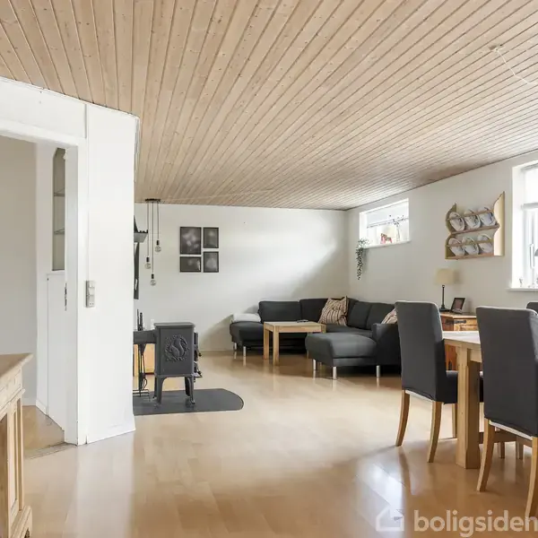 Stue med grå sofa og sofabord, siddende til venstre. Spisesæt med træbord og seks stole til højre. Lyse trægulve og loft. Hylder og billeder på væggene. Store vinduer.