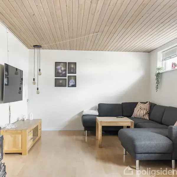En sort sofa er placeret i et lyst stuehjørne med puder, et sofabord i træ, ophængte lamper, en væghængt fladskærm og billeder. Rummet har trægulv og loft.