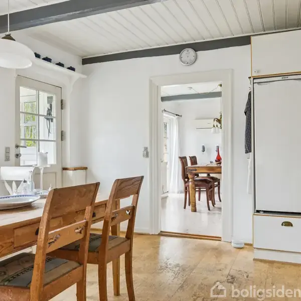 Et køkken med spisebord og stole står i forgrunden. Der er en dør med glasruder i baggrunden og en stor hvid køleskab ved siden af en indbygget ovn. En åbning leder til et tilstødende rum.