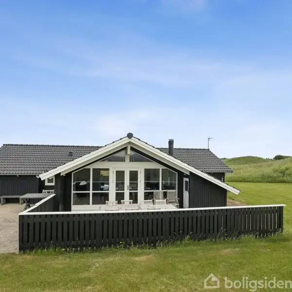 Sort hus med store vinduer står stille omgivet af en grøn græsplæne og blå himmel. Terrasse med sort hegn foran huset.