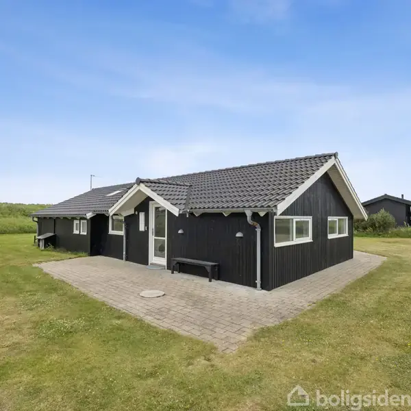 Sort træhus står på en velplejet græsplæne. Huset har hvide vinduesrammer, en grå tagryg og en flisebelagt terrasse foran. Omgivelserne består af grønt landskab.