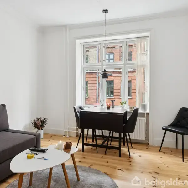 Sofa står i en lys stue ved siden af et lille, hvidt sofabord. Et spisebord med fire stole er placeret ved vinduet, og en stol står ved siden af.
