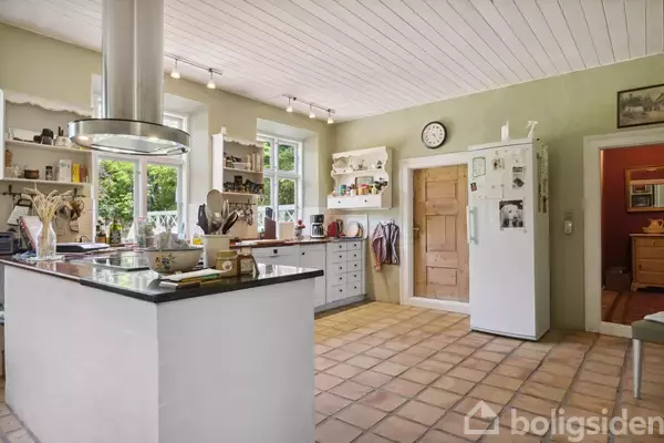 Et køkken med en kogeø i midten, omgivet af skabe og hylder. Køleskabet står ved siden af en trædør. Væggene er lysegrønne, og gulvet er flisebelagt. En væg ur hænger over køleskabet.