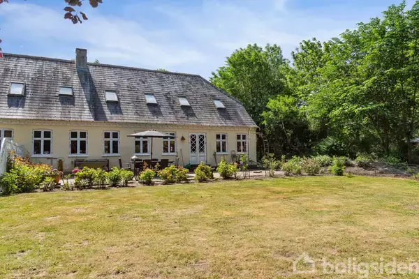 Et gult hus med skråt tag og flere vinduer står i en have med velplejet græsplæne. En parasol og havemøbler er arrangeret foran huset. Skov i baggrunden.