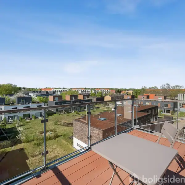 En balkon møbleret med to stole og et bord står stille, omgivet af en glasafskærmning. Udsigt over et moderne boligområde med grønne arealer og flade tage under en klar blå himmel.