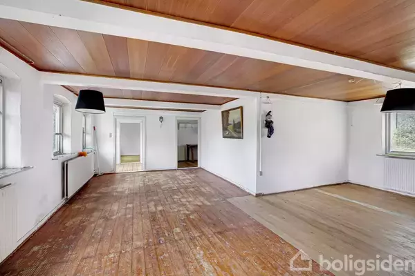 Et rummeligt rum med træloft og trægulv, ingen møbler, to sorte lamper hænger fra loftet, hvide vægge, flere vinduer, en lille radiator og en dør i baggrunden.
