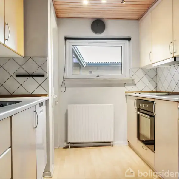 En spisebordsgruppe med seks stole står i en stue, hvor et sort lænestol og et fjernsyn også ses. Rummet har lyse vægge og store vinduer med udsigt til en terrasse.