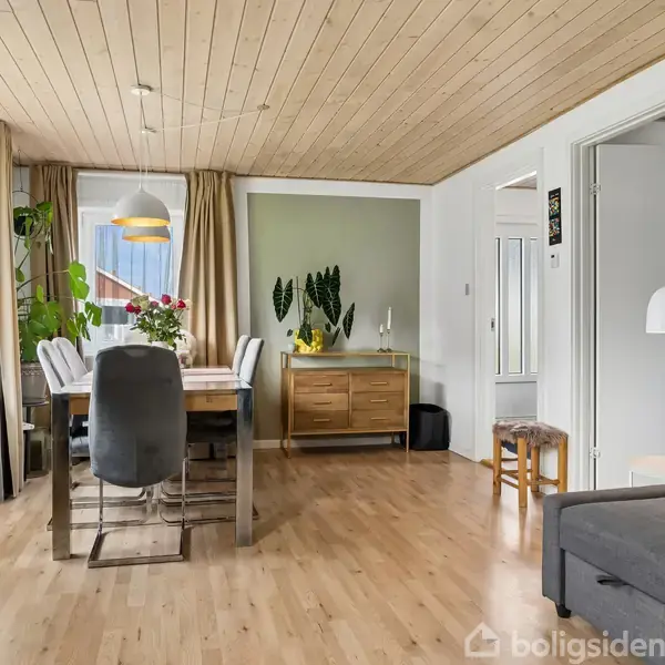 Spisebord med stole står i en stue med lyst trægulv og loft. Bordet er dækket med planter og en lampe hænger over. En kommode står ved væggen.
