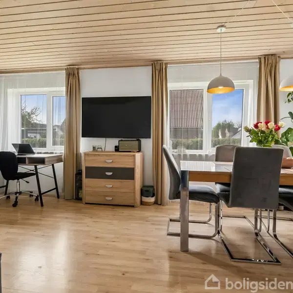 Stue med trægulve og -loft. Bord og stole ved vindue med gardiner. Fladskærm på kommode. Skrivebord med stol ved siden af. Plante og vase med blomster på bordet.