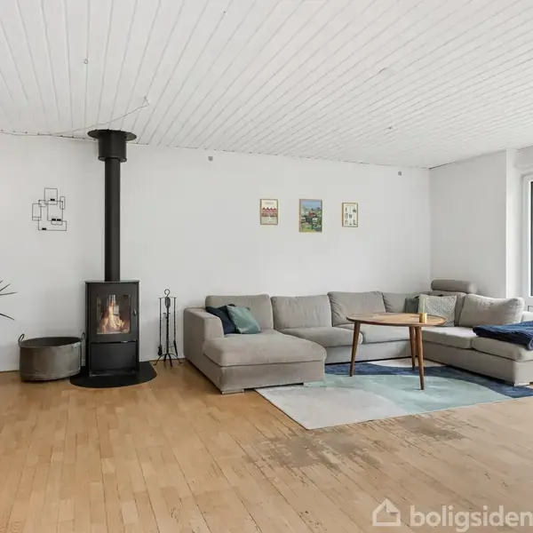 En moderne stue med en grå hjørnesofa og et træbord udfolder sig, mens en brændeovn står tændt. Et stort vindue giver udsigt til en grøn have.