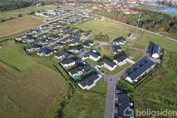 Arealfoto af en række nye parcelhuse med mørke tage, arrangeret i en bølgende formation. Husene omgives af grønne græsarealer og veje, med en skov i baggrunden.