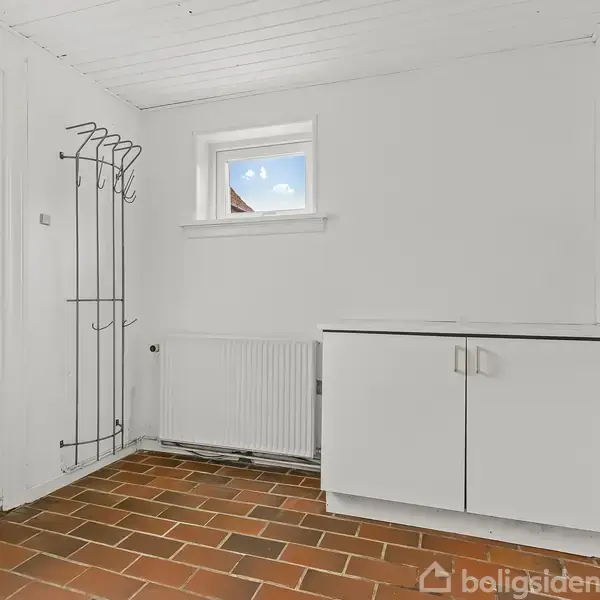 Et opbevaringsrum med en hvid skabsenhed står langs en væg. En dør til venstre, en knagerække, og et vindue ovenover en radiator supplerer rummet med murstensgulv.