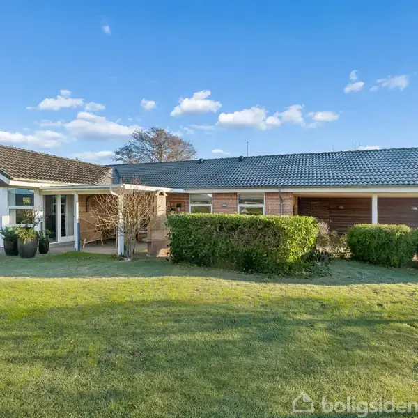 Et etplanshus med rødt murværk og gråt tag står stille i en solrig have, omgivet af grønne buske og velplejet græsplæne under en klar, blå himmel.