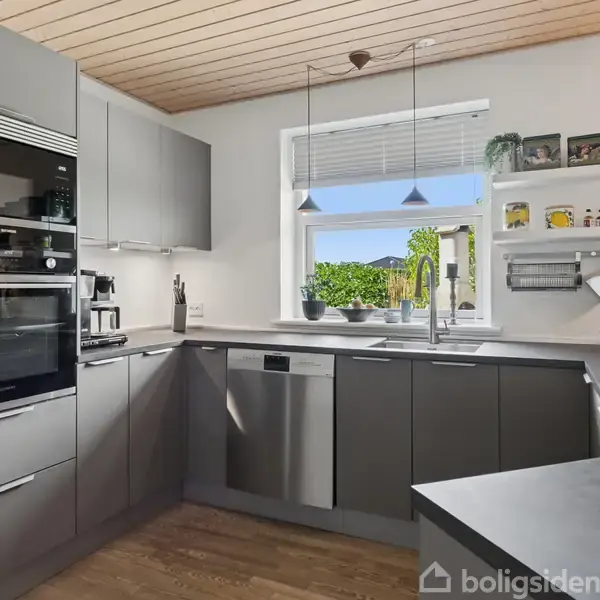 Et moderne køkken med grå skabslåger og indbyggede apparater. En opvaskemaskine og vask ses foran et vindue med udsigt til en have. En kaffemaskine står på bordet.