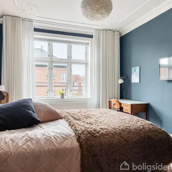 Soveværelse med dobbeltseng, pyntepuder og tæppe; vægmonteret fjernsyn over skrivebord med lampe. Store vinduer med hvide gardiner, blå vægge og elegant stukatur i loftet.