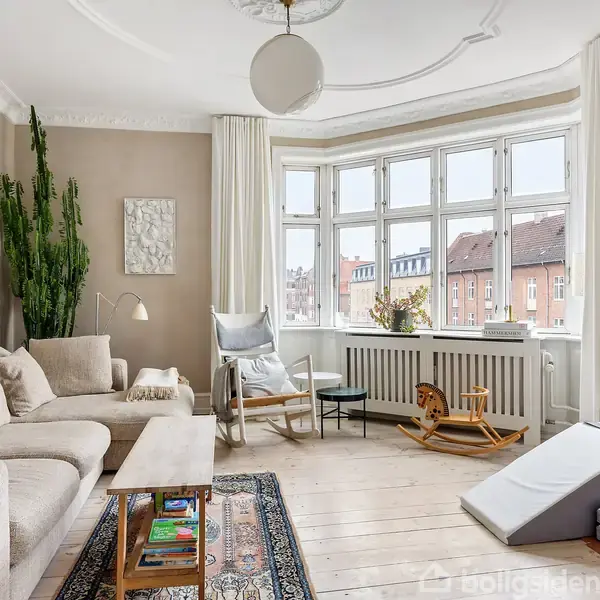 En lys stue med en beige sofa og tæppe. En stor kaktus står i hjørnet. På gulvet ligger et tæppe, og der er legetøj nær vinduet med udsigt over byen.