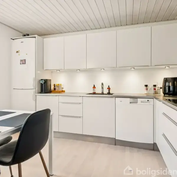 Et moderne køkken indeholder hvide skabe, en opvaskemaskine, og sort elektronik på bordpladen. En spisekrog med sort bord og stole er til venstre, og der er masser af naturligt lys.