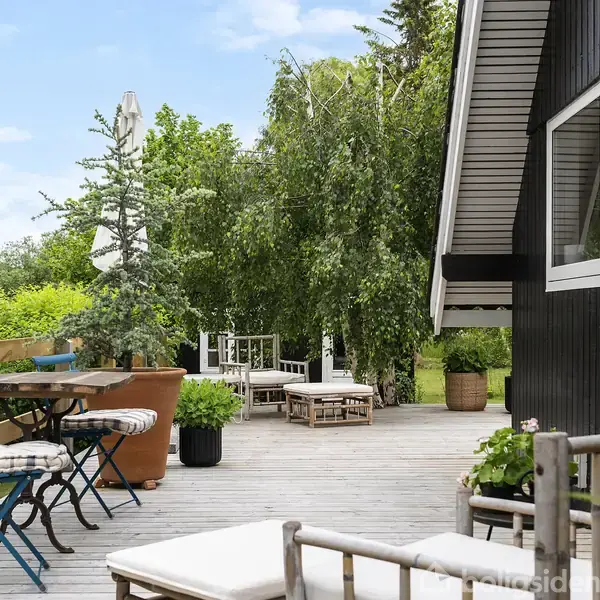 Terrasse med sortmalet husvæg har blå og hvide caféborde og stole, omgivet af frodig have med træer og planter. Solrigt, afslappet udendørsområde ideelt til ophold.