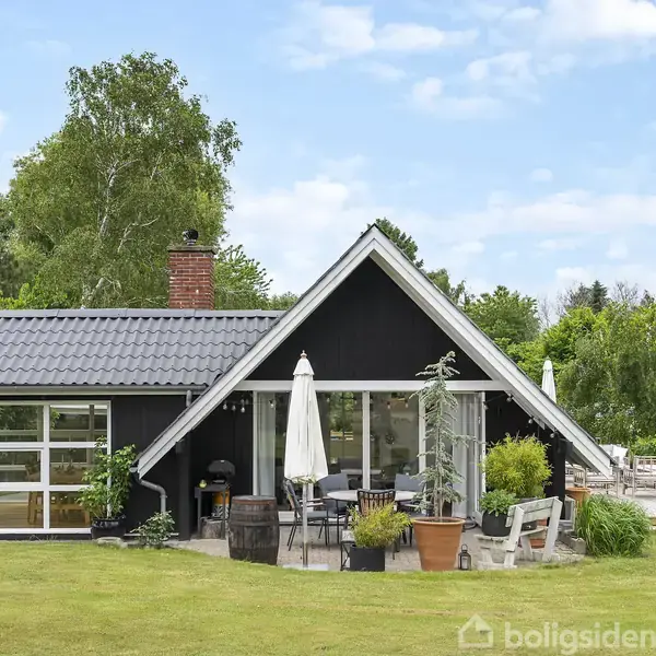Sort hus med skrånende tag, terrasse og have; omgivet af grønne træer og buske. Havemøbler på terrassen med en parasol i midten. Græsplæne foran huset.