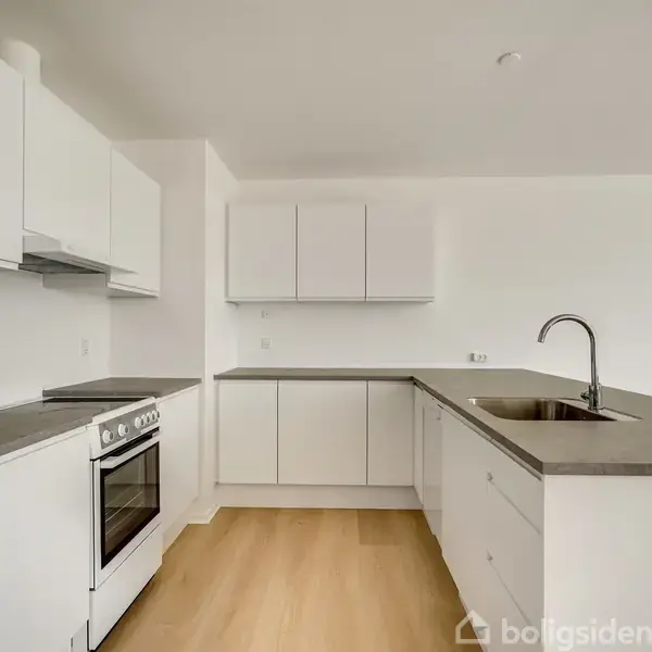 Hvidt køkken med moderne, integrerede skabe og skuffer. Komfur og vask er installeret, og der er trælaminatgulv. Væggene er hvide, hvilket giver et minimalistisk udtryk.