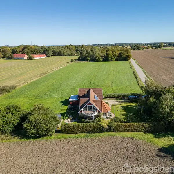Et hus står midt i en grøn mark omgivet af tæt, lav vegetation. I baggrunden ses flere åbne marker og træer under en klar blå himmel.