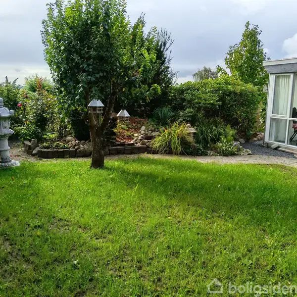 Have står stille i en frodig have med græsplæne og buske. Til venstre ses en stenskulptur; en glaspavillon står til højre.