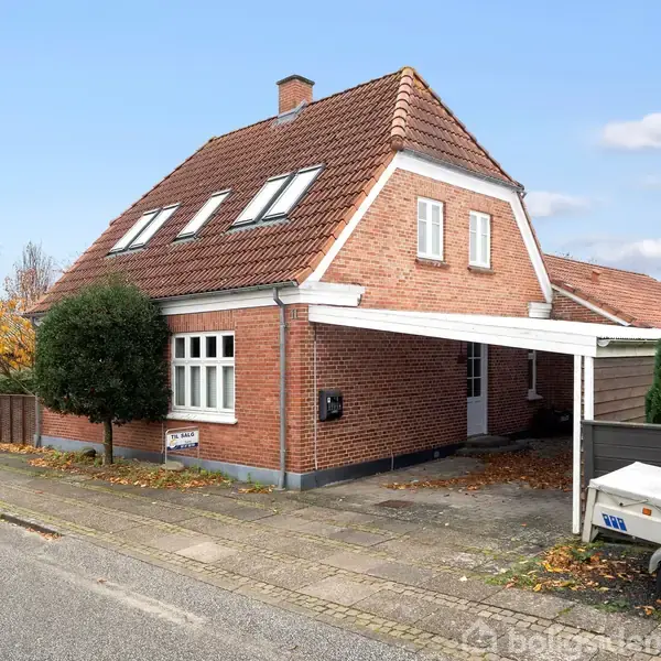 Et rødt murstenshus med skråt tag har en carport ved siden af. Foran er en trailer parkeret på fortovet. Omgivelserne er et villakvarter med træer og et fortov.