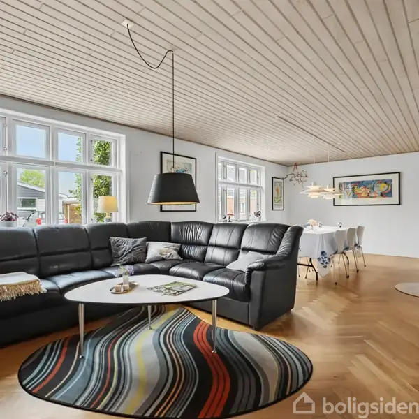 Sort hjørnesofa står i en stue, dekoreret med farverigt tæppe nedenunder. Et rundt sofabord er foran sofaen, og rummet har trægulv og hvide vægge med vinduer.