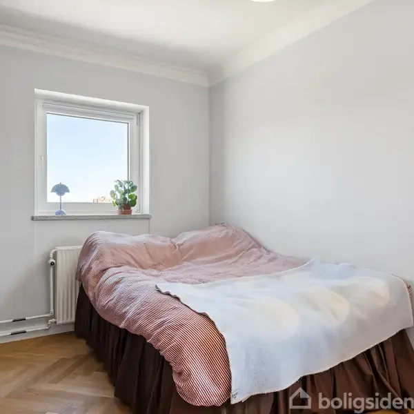 Sengen er redt med lyseblåt og rødt sengetøj, placeret ved vindue med plante og lampe på vindueskarmen. Skabet står til venstre. Rummet har lys væg og parketgulv.