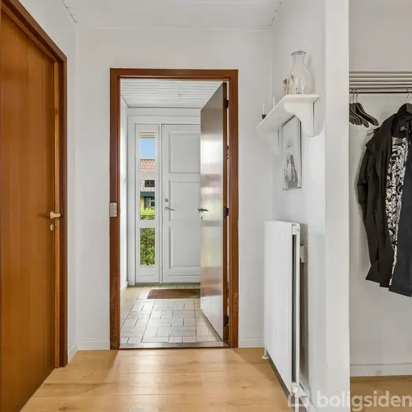 En gang med træplankegulv fører til en hvid hoveddør. Venstre side har en lukket, brun dør og billede på væggen. Højre side har en åben garderobe med jakker.